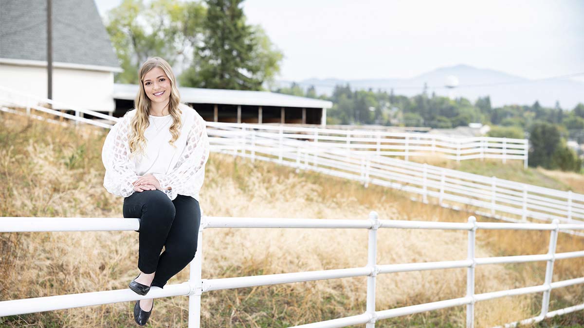 A woman sits on a white fence with a white barn in the background.