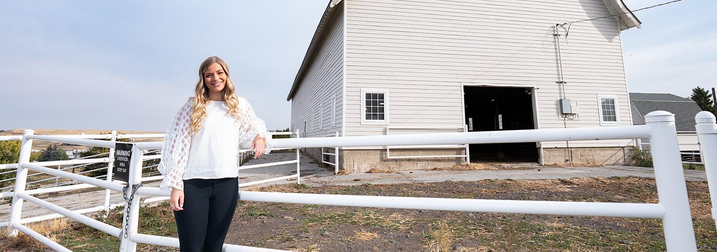 A woman stands next to a white fence with a white barn in the background.