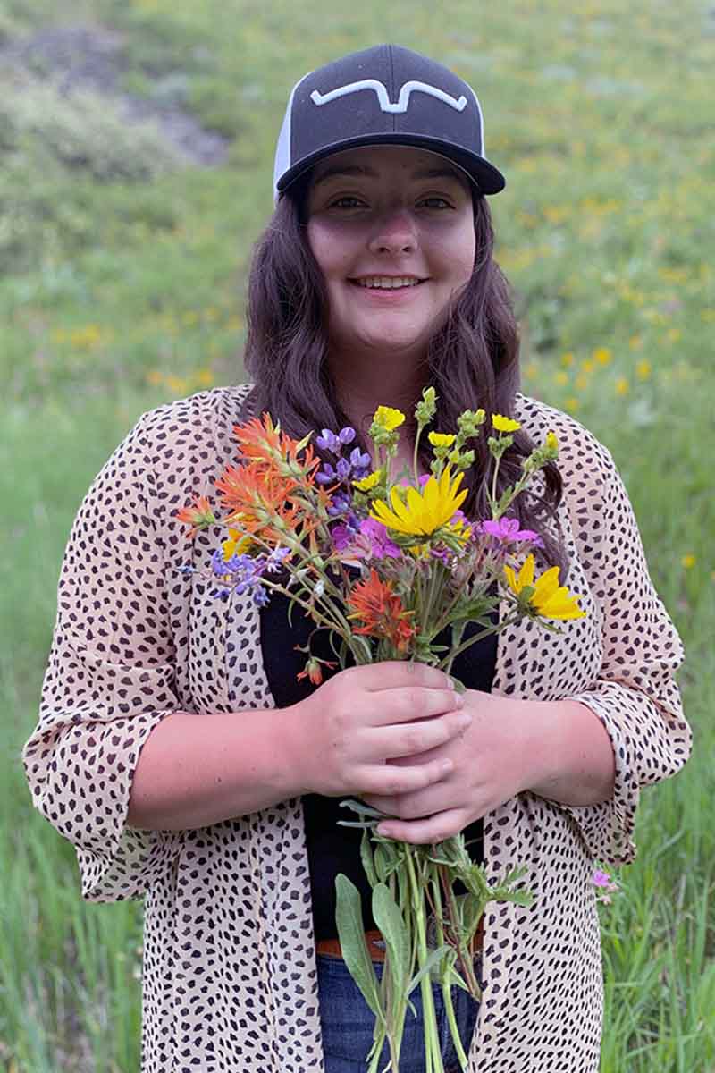 A young woman wearing a hat, holding flowers in a field.