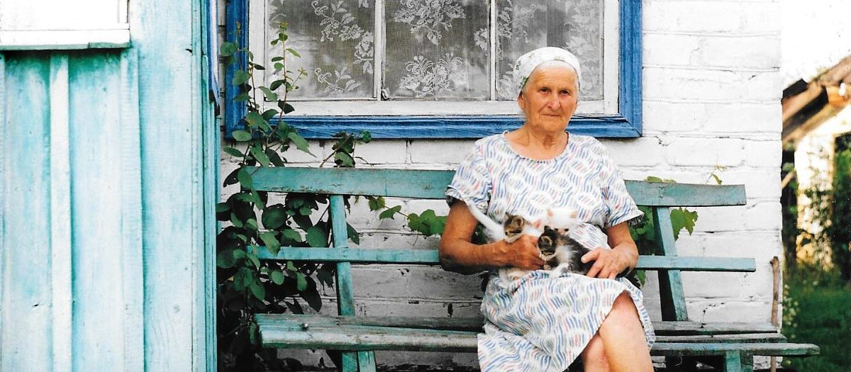 An elderly woman sitting on a bench with three kittens in her lap outside her house in the Ukraine.