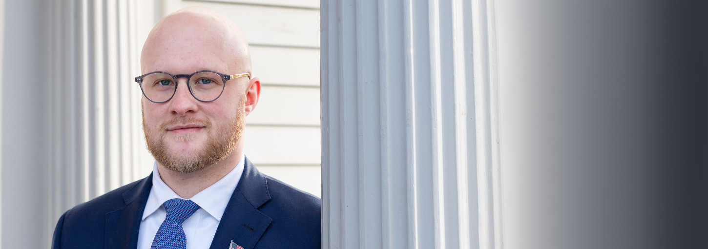 A bearded, bald man with eyeglasses, wearing a suit with an American flag lapel pin stands near a Doric column.