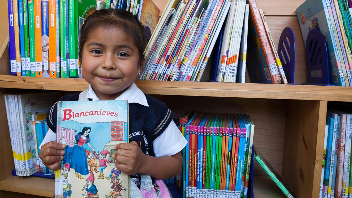 A child stands holds a copy of "Blancanieves," Snow White in Spanish, in front of a library shelf at her school.