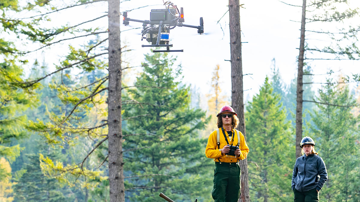 A drone lifts off in a forest in front of the drone pilot who wears yellow firefighting clothing.