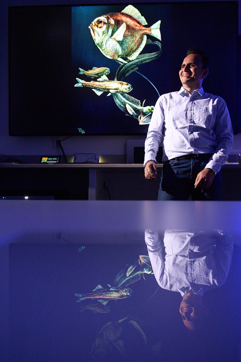 Jagdish Patel stands in front of a projection of three deep-sea fish.