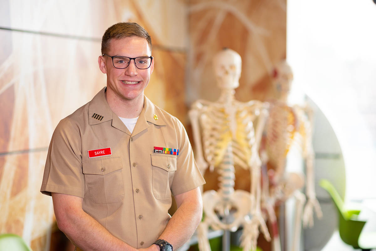 Young man in military service blouse and slacks stands in front of two model human skeletons.