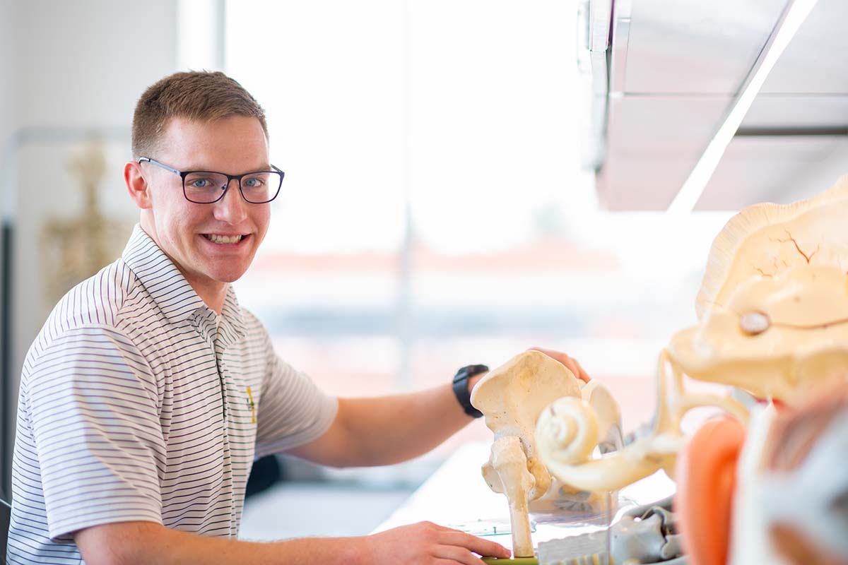 Young man in sport shirt holds a hip bone model while sitting at a desk.