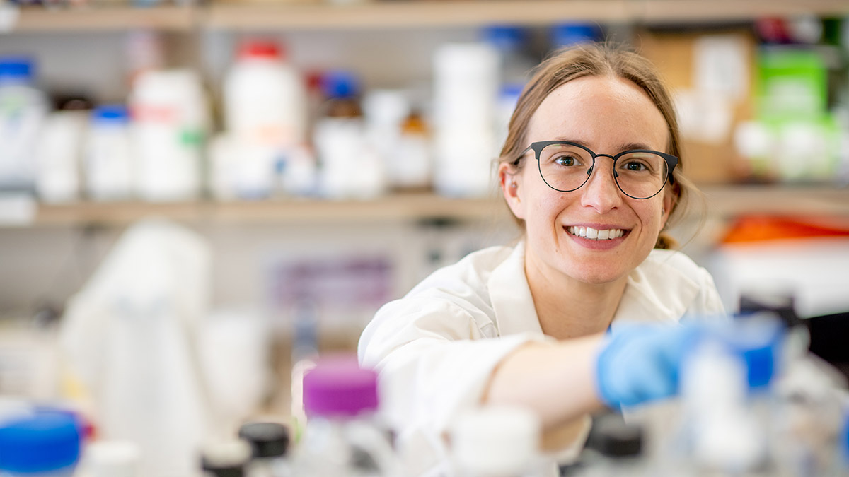 Female scientist smiles from behind beakers and chemistry glassware while seated in a chemistry lab.