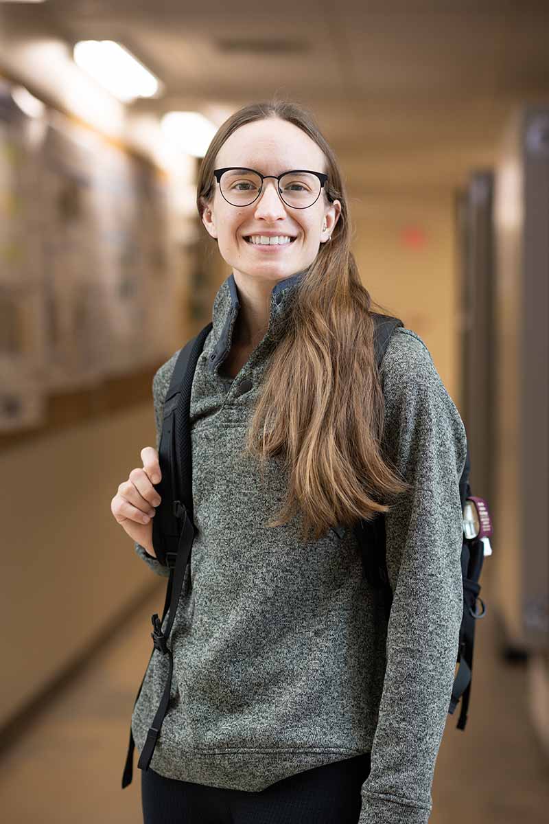 Smiling female college student with long hair and glasses stands in hallway with a backpack.