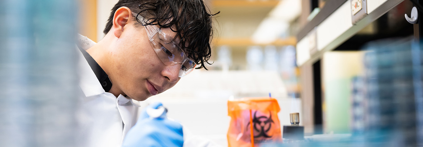 Scientist wearing safety goggles, lab coat and blue gloves uses pipette to drip liquid into a Petri dish.