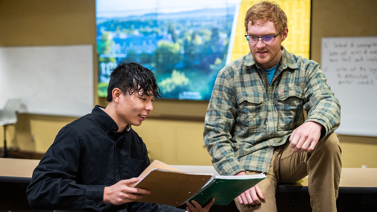 Two students, one seated on a chair holding an open binder, and another seated nearby on a table, discuss a topic in the open binder.