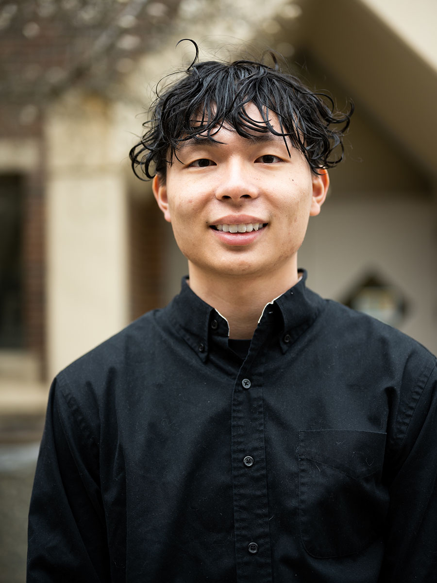 A student with dark hair and a dark shirt stands in front of a brick building.