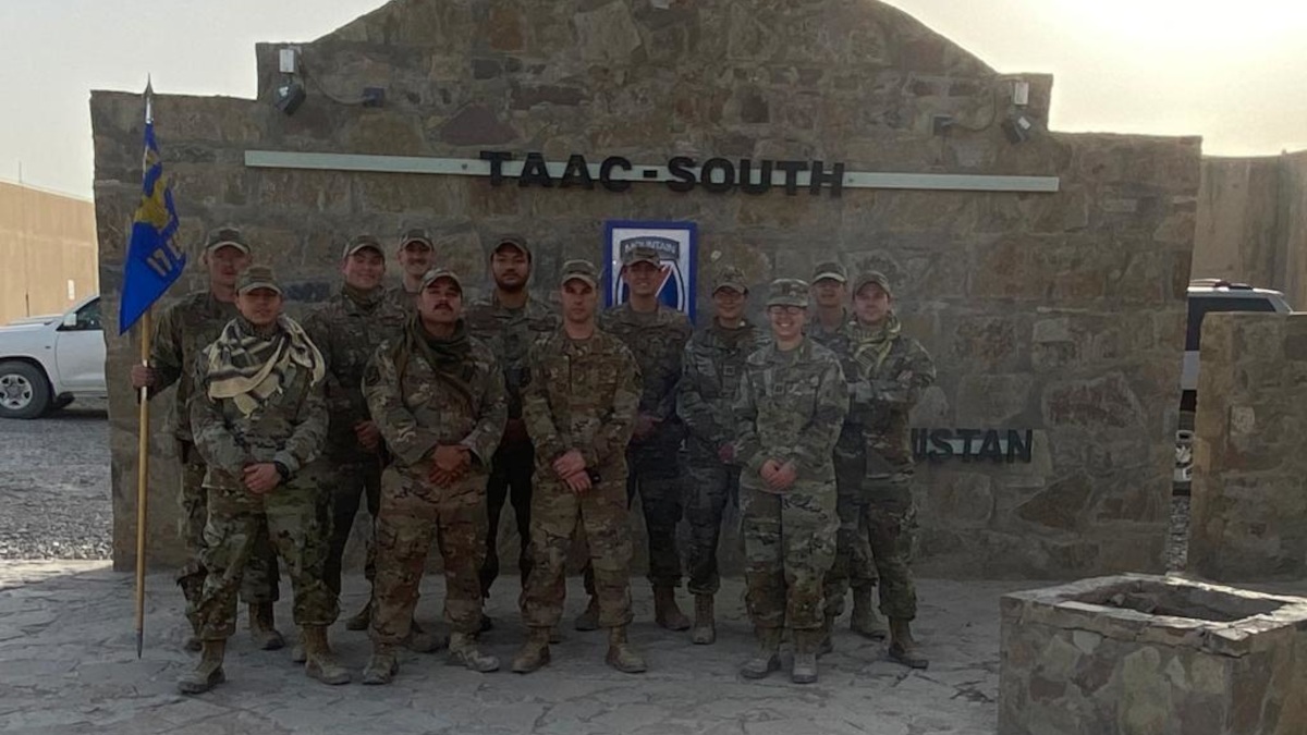 Eugene Russell stands with a group of eleven other team members in military gear in front of a landmark sign for TAAC South.