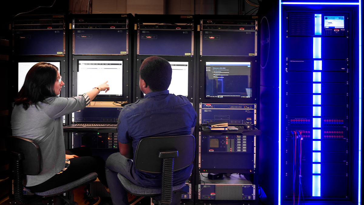 Two computer scientists discuss the information displayed on a screen next to a large server rack.