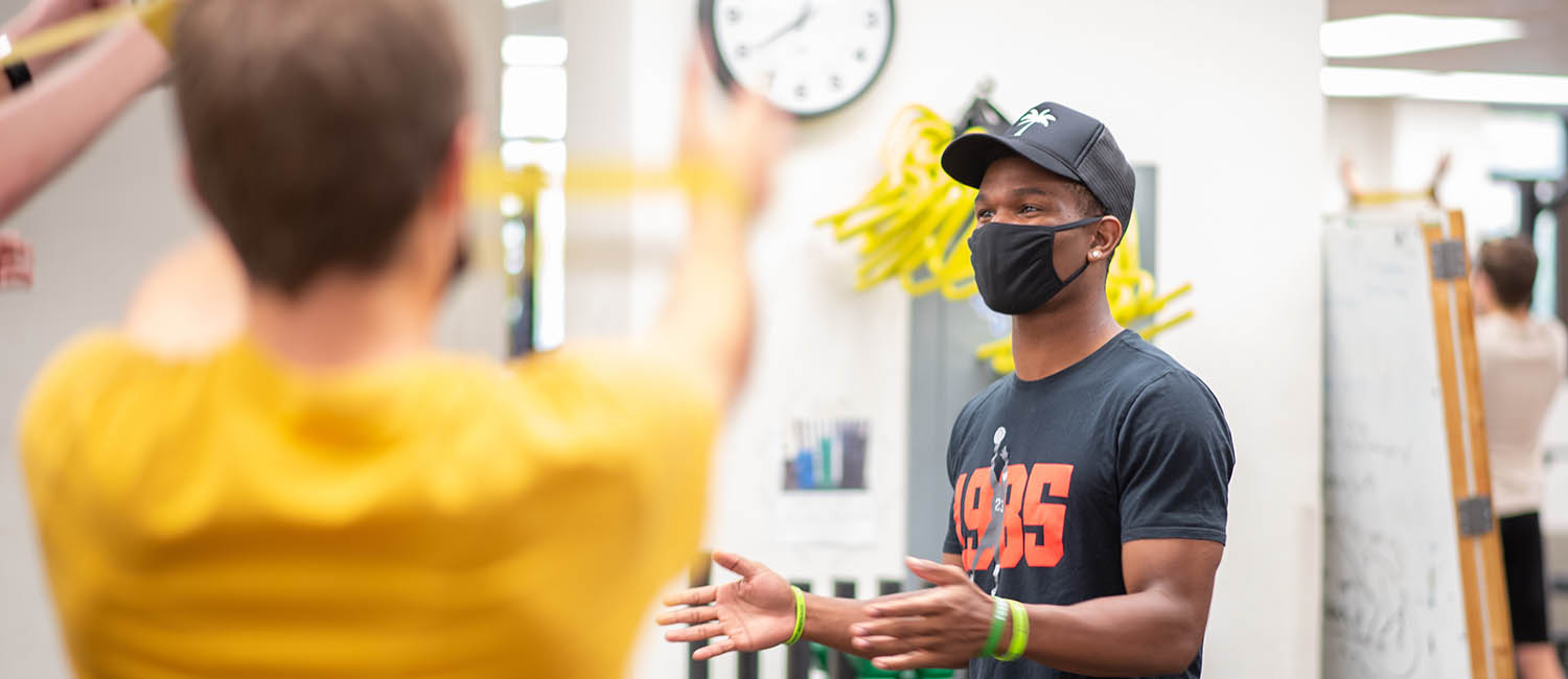 University of Idaho student Wyryor Noil, pronounced warrior, faces to left of frame and smiles under black mask while demonstrating proper arm placement to Moscow High School PE student wearing bright gold shirt in near foreground.