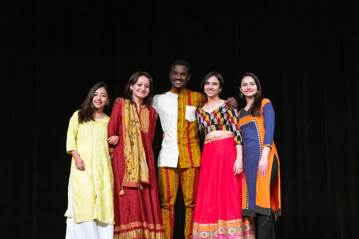 A group of people wearing clothes from various cultures pose in front of a black curtain