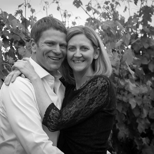 Owner Denise Isenhower smiles while wrapped around Brett Isenhower, both sharing in each other's happiness with grape vines in the background.