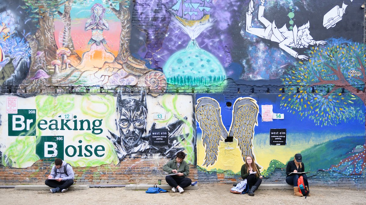 Four students sit on the ground sketching in front of a wall painted with colorful murals. One reads "Breaking Boise."