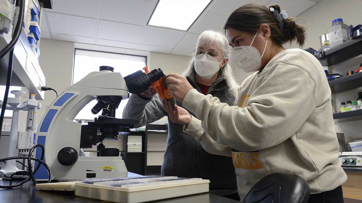 Two women inspect a microscope in a lab.