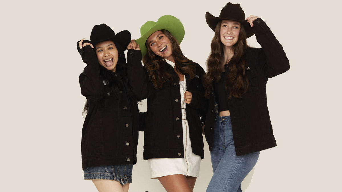 Three women wearing coats and cowboy hats smile at the camera.
