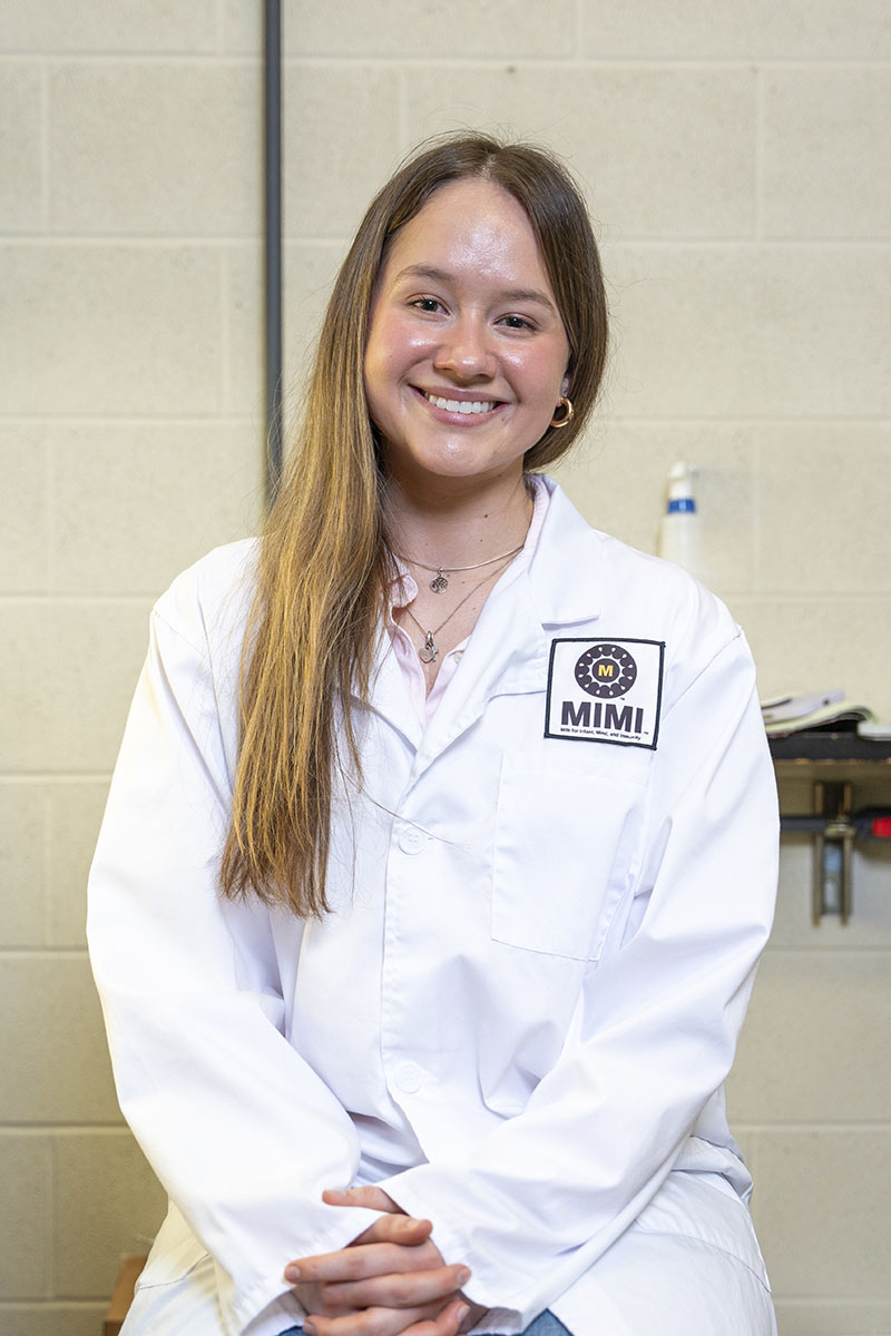Female researcher sitting on a chair in the Milk for Infant, Mind and Immunity (MIMI) Laboratory, smiling for the camera in her MIMI lab coat.