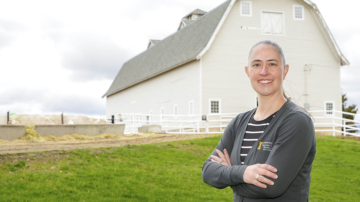 A woman standing in grass field with a tan barn and fencing in the background.