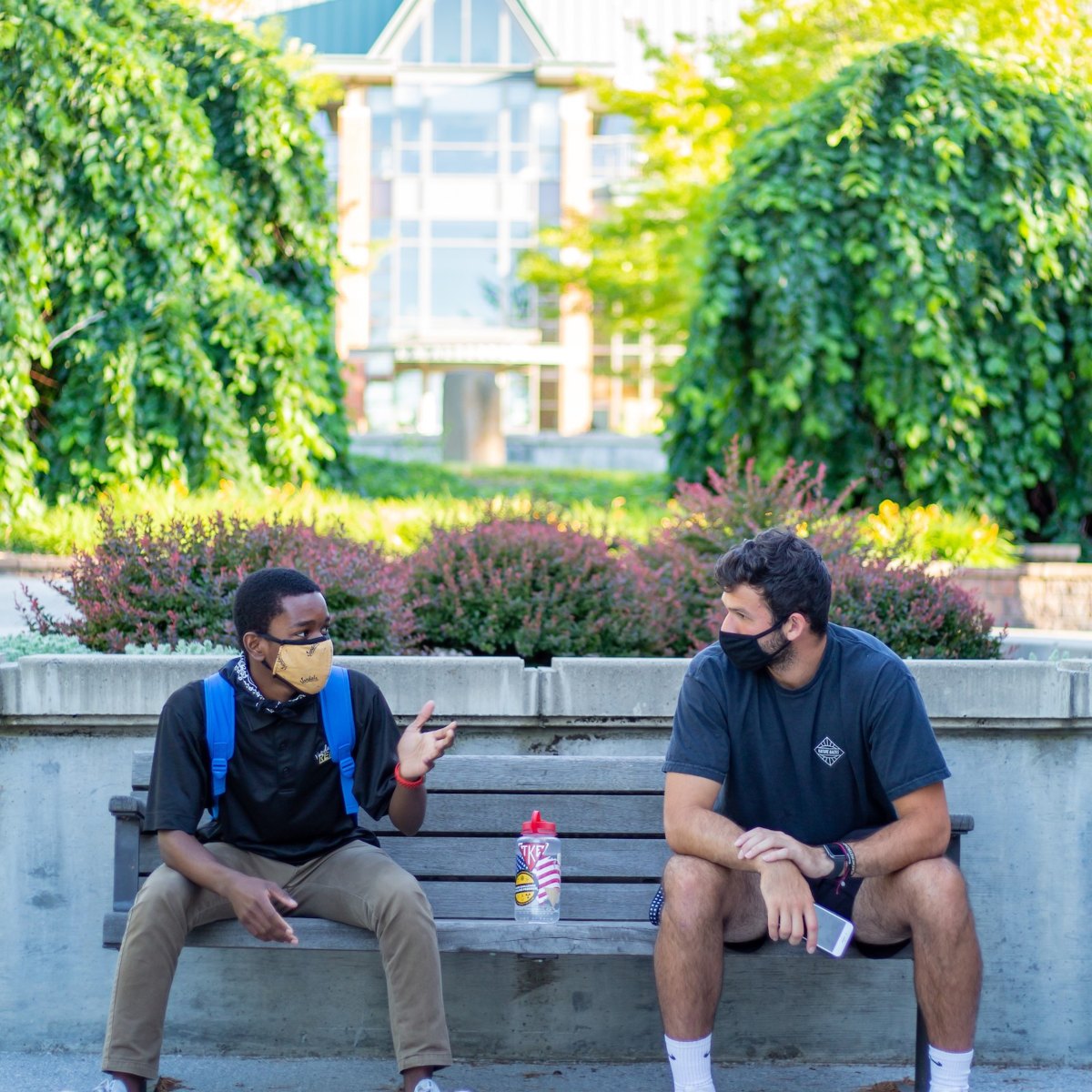 Two students wearing Vandal-themed face coverings enjoy a conversation on a bench outside the J.A. Albertson building.