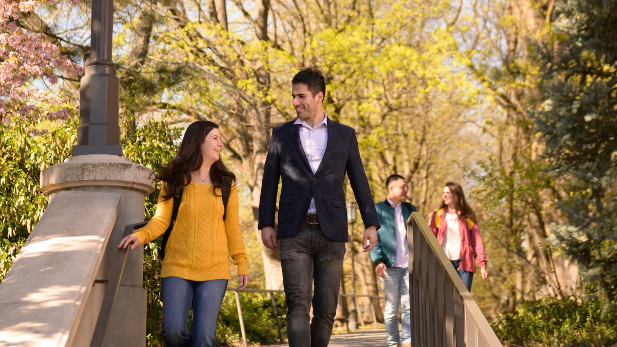 A female in jeans and a gold sweater talks with a male in a blazer as they walk down Hello Walk on the University of Idaho campus.