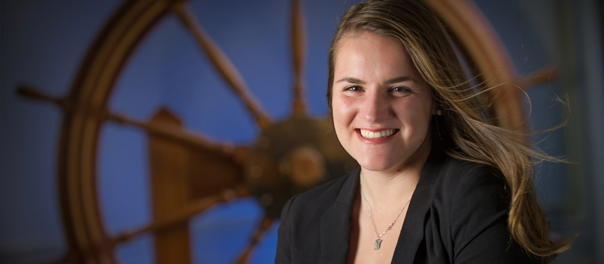 A student poses in front of a ship's steering wheel.