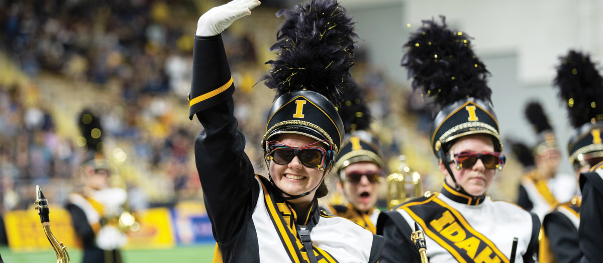 A female marching band member waves up at the fans while performing on the football field. 