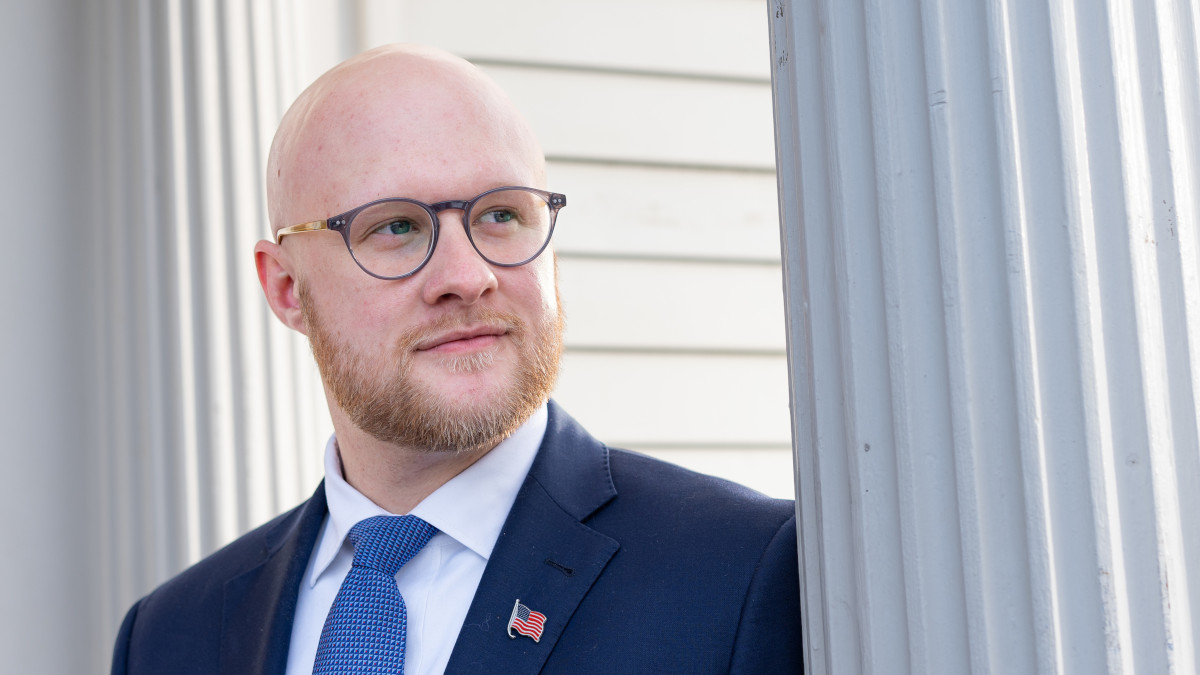 A bearded, bald man with eyeglasses, wearing a suit with an American flag lapel pin stands near a Doric column.