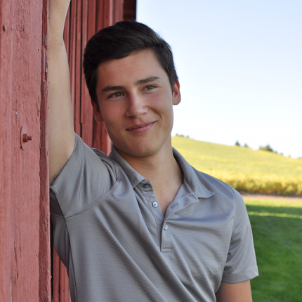 Jayson Kristopher Gray leans against the wooden side of a building in the shade, green fields behind him.
