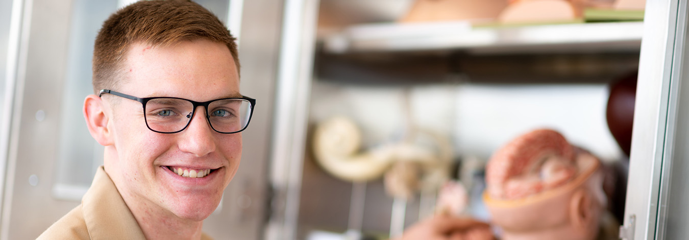 Young man wearing glasses, military dress shirt, with one hand in a cupboard of medical anatomy models.