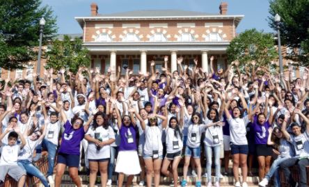 Hundreds of teens raise their hands in a cheering gesture. Most wear white or purple t-shirts and shorts. They stand outdoors in front of a building with columns.
