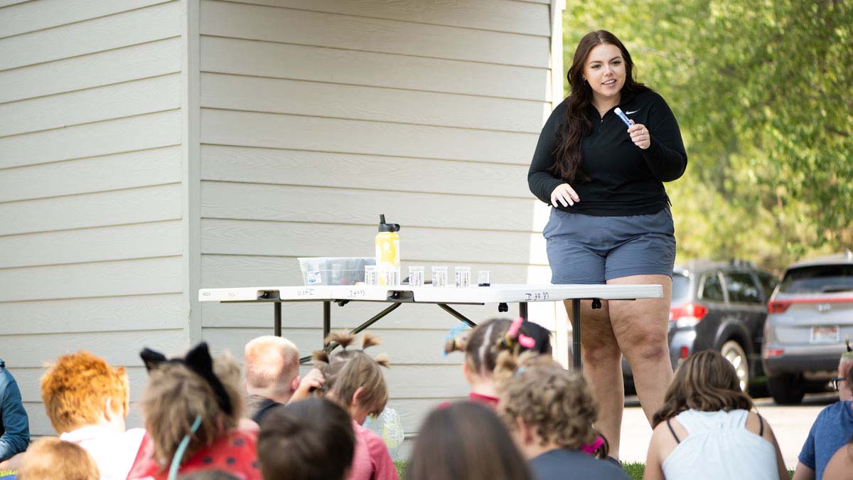 Emma Arman stands behind a table outside, talking to a group of young campers all with their backs to the camera. 
