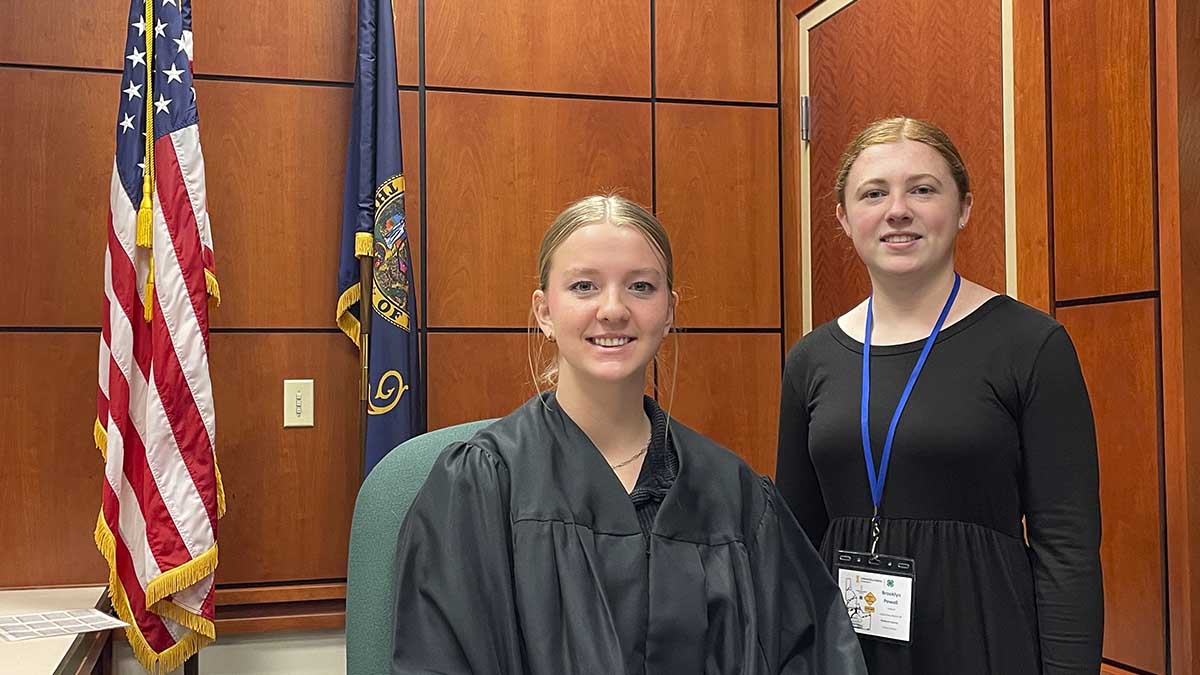 A young woman wearing a judges robe sits while another young woman stands next to her.