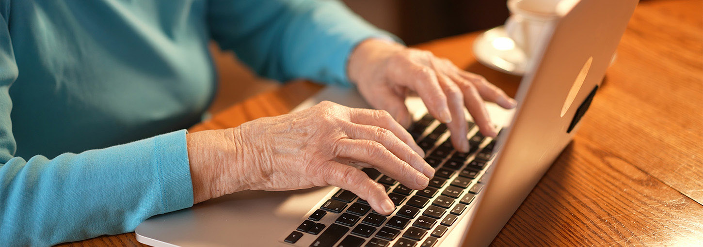 An elderly woman’s hands rest on the keyboard of a laptop.