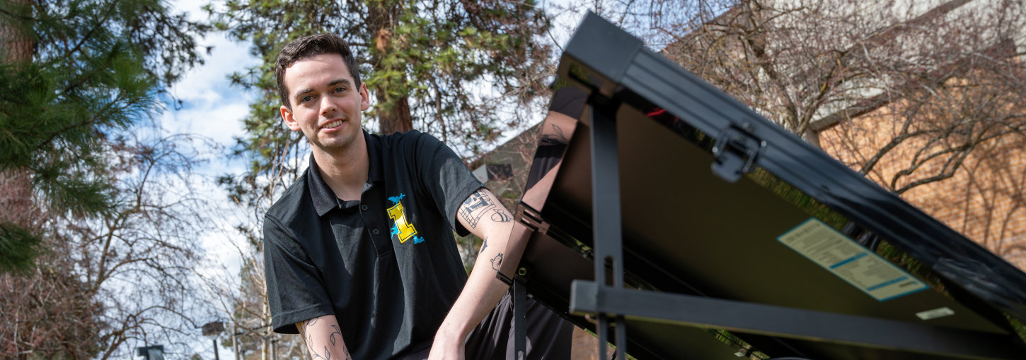 A U of I Coeur d’Alene student smiles while kneeling in the grass while working on equipment. 