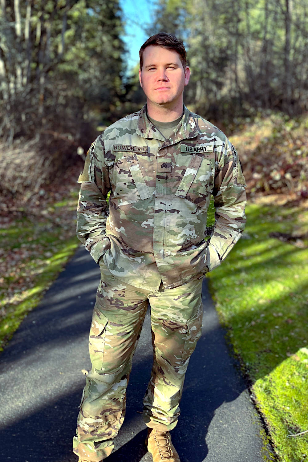 Man in battle dress uniform stands on a paved park trail.