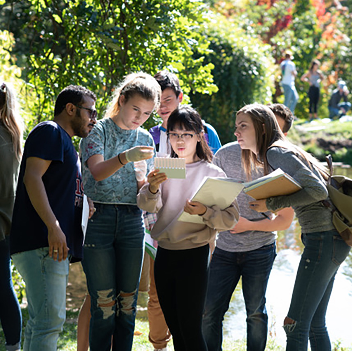 Students with notepads stand in front of a pond.