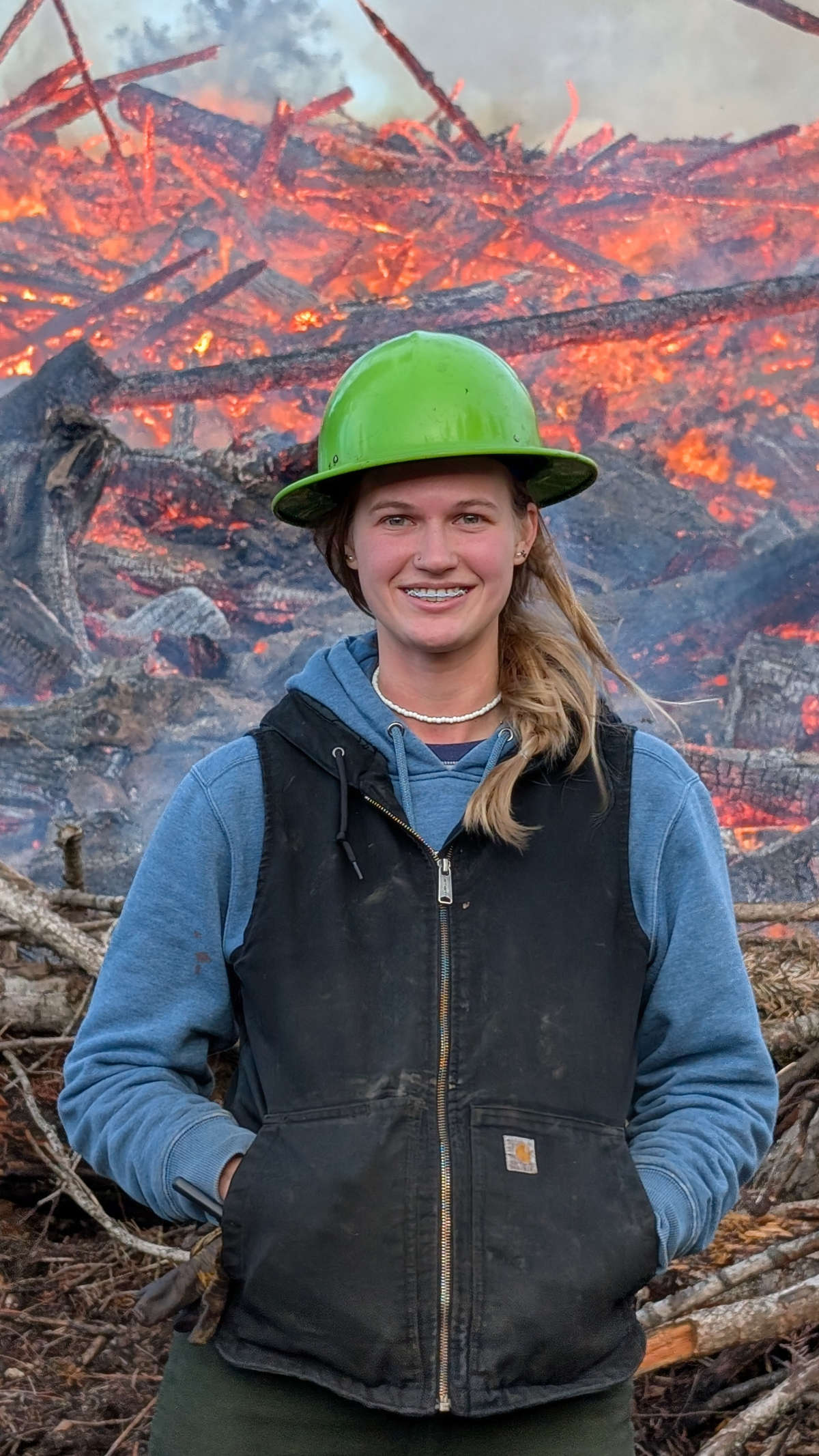 Woman with hardhat stands in front of a slash fire.