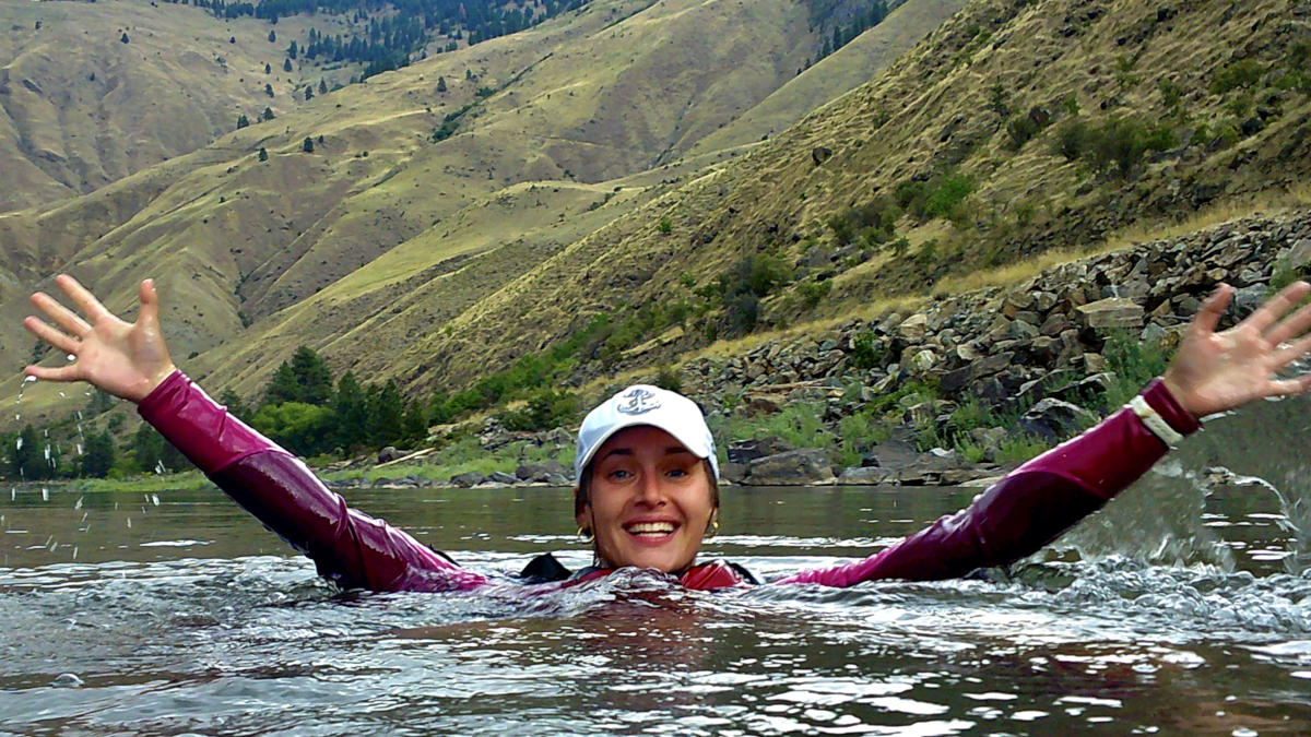 Woman wearing a ballcap raises arms while swimming in a river.