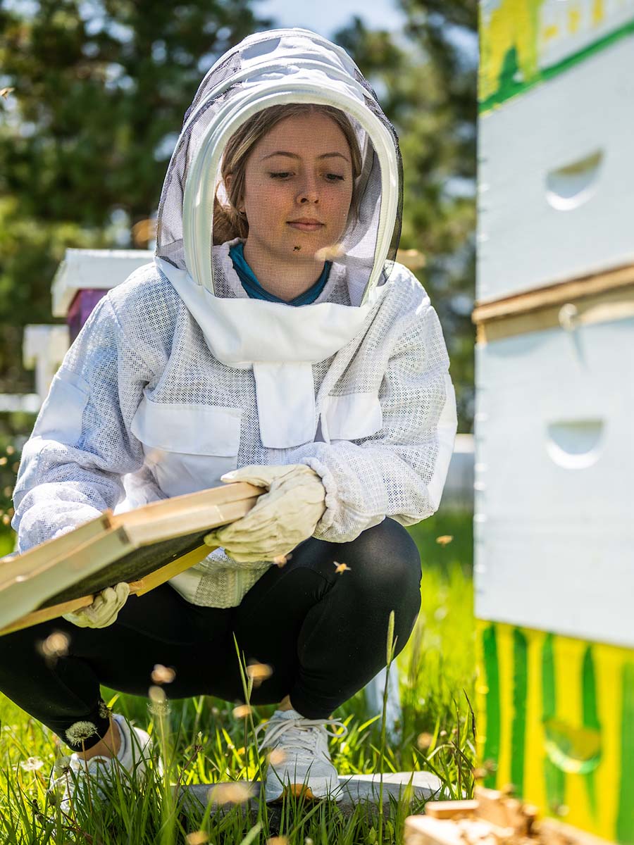 woman in bee suit kneels in grass in front of beehive.