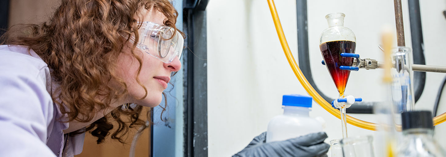 Woman in chemistry lab wearing safety goggles and lab coat examines a beaker.