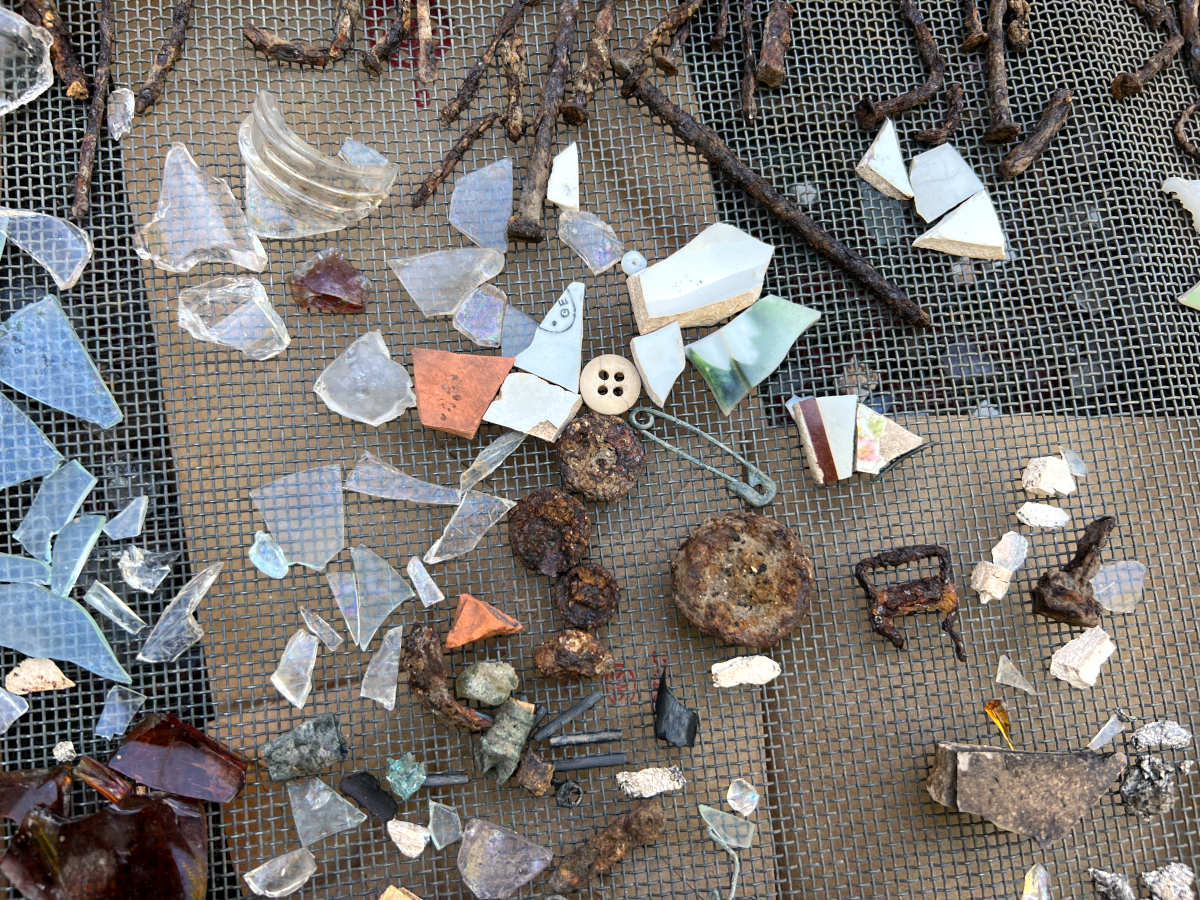 A large metal mesh screen holds pieces of a comb, shards of bottle glass, buttons and coins.