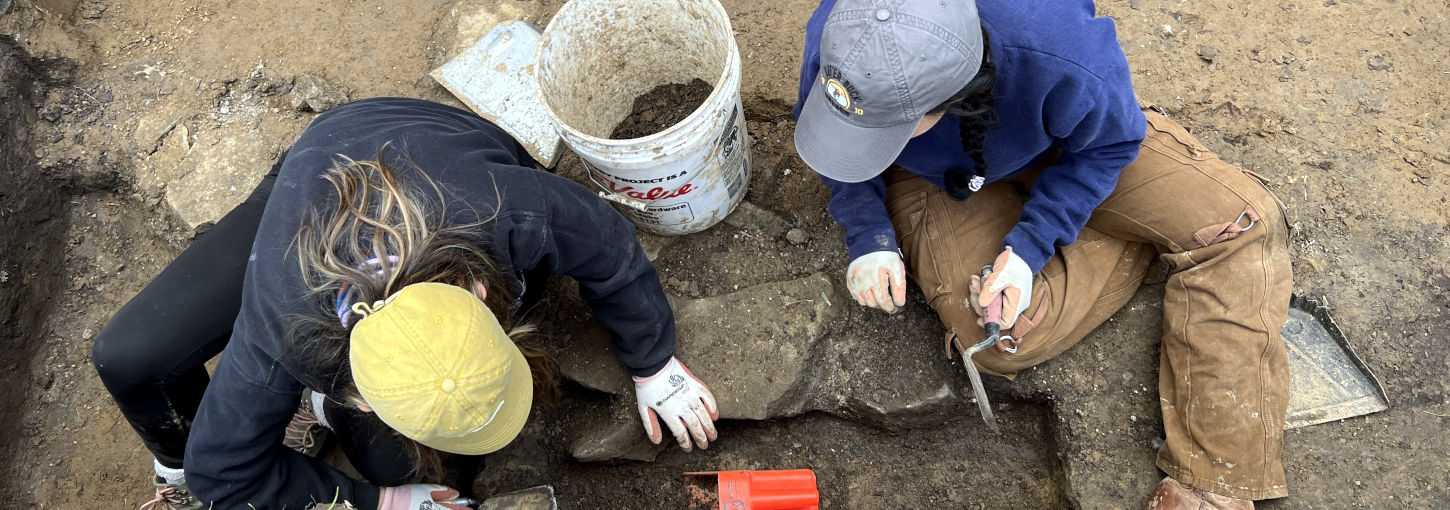 Two students use trowels as they dig dirt in a rectangular pit.
