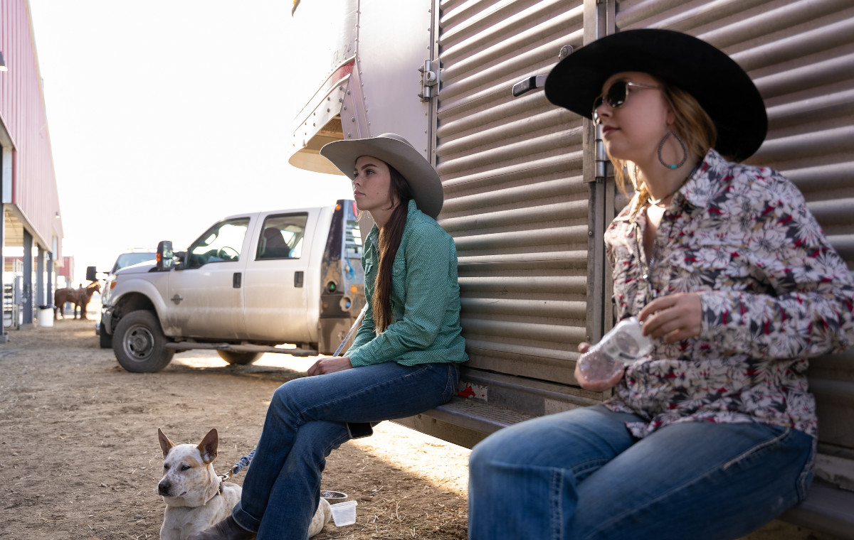 Two women in cowgirl hats sit on the back of a horse trailer.