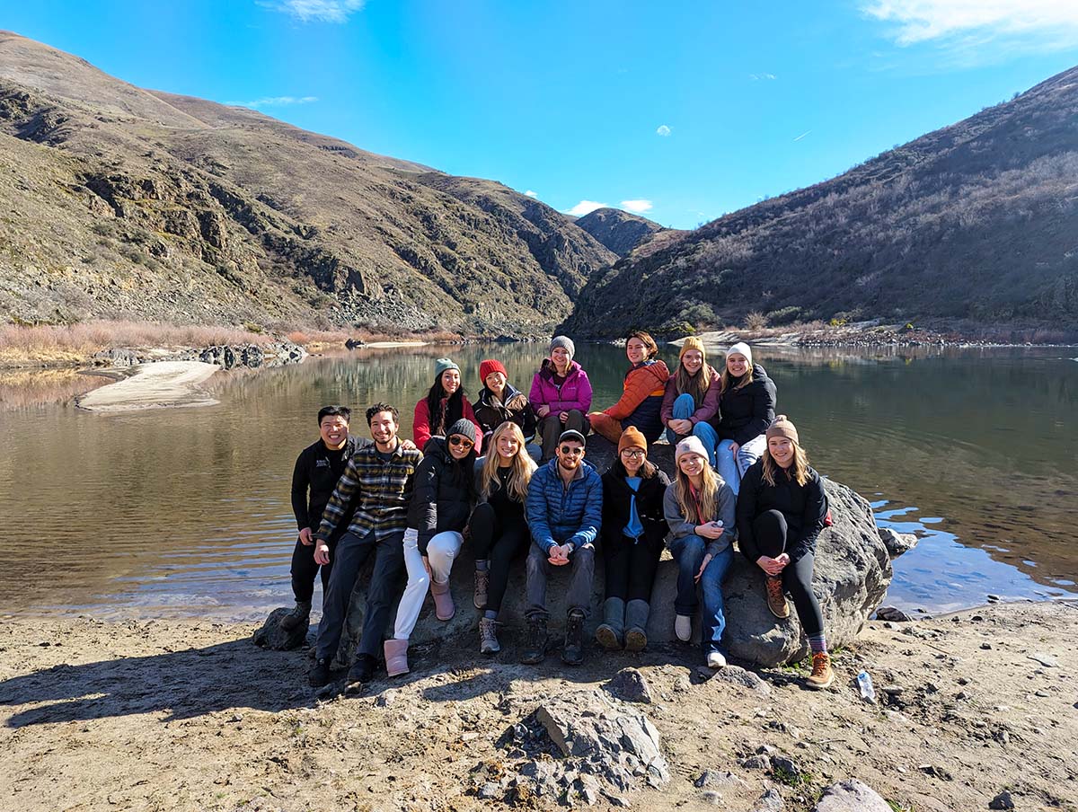 A group of medical students sit in front of a lake and mountains.