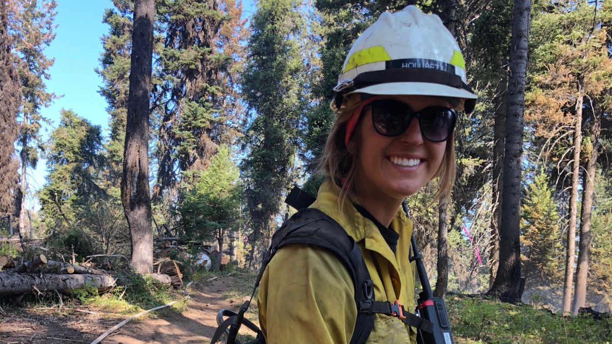 Heidi Holubetz stands in a forest in yellow and green firefighting gear.