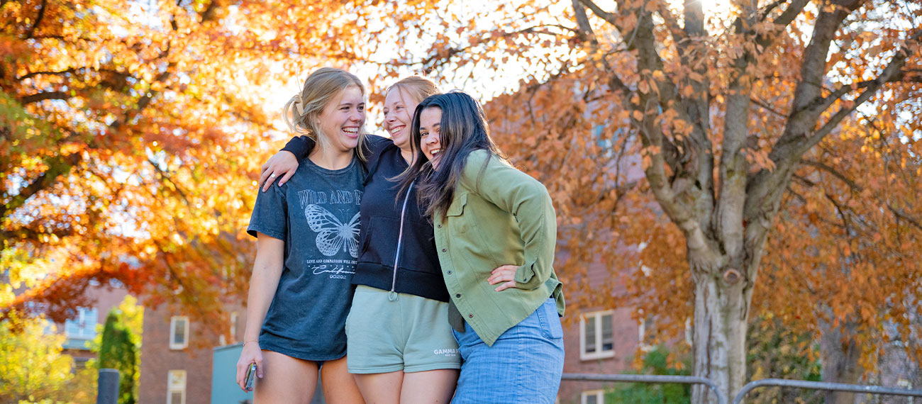 three students hugging, standing in front of fall colored trees