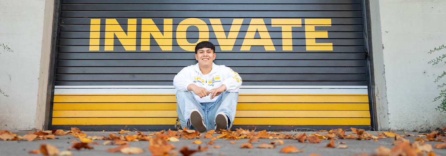 A University of Idaho student sits outside on the ground in front of a sign that says innovate.